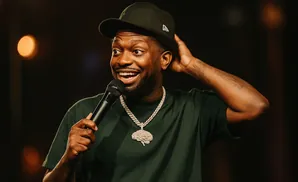 Comedian wearing a green shirt and hat, holding a microphone and touching his head.