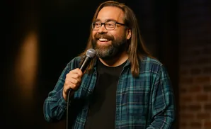 A man with long brown hair, a beard, and glasses holds a microphone while wearing a blue plaid shirt.