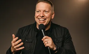 A man in a black jacket smiles and speaks into a microphone with his left hand raised.