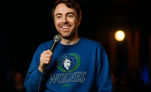 A man in a blue sweatshirt with a wolf logo grins while holding a microphone.