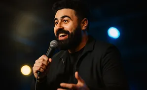A man with a beard speaks into a microphone on a dimly lit stage.