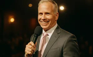 A man in a suit and tie smiles while holding a microphone.