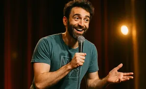 A smiling man with a beard holds a microphone, gesturing with his other hand.