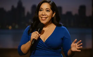 A smiling woman in a blue dress holds a microphone onstage with a cityscape in the background.