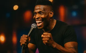 A Black man with a stylish haircut laughs while holding a microphone, his fists clenched.