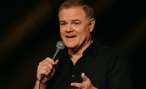 A man in a black shirt holds a microphone while speaking and gesturing.