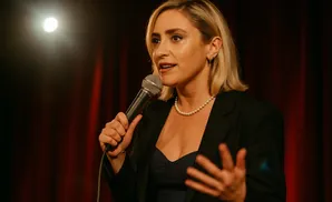 Woman with blonde hair speaks into a microphone, wearing a black blazer and pearl necklace.