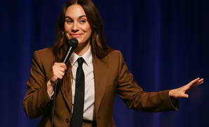 A woman in a brown suit and tie holds a microphone and gestures to the right.