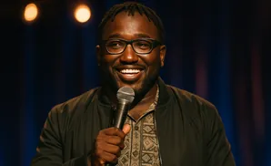 A Black man with dreadlocks and glasses smiles while holding a microphone and gesturing with his hands.