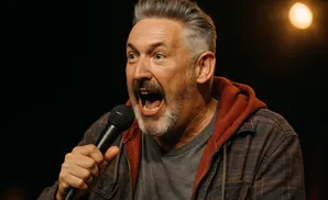 A man with grey hair and beard, wearing a plaid jacket, shouts into a microphone.