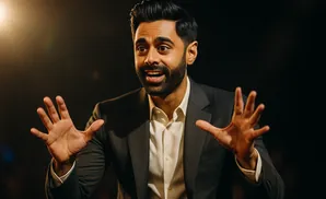 A man in a suit gestures with both hands, speaking animatedly in front of a dark background.