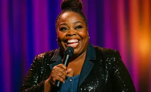 A Black woman with her hair in a bun, wearing a sequined jacket, laughs while holding a microphone onstage.