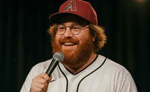 A bearded man in a baseball cap and jersey holds a microphone and smiles.