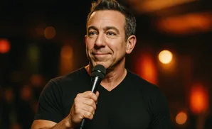 Comedian holding a microphone and smiling, with warm bokeh lights in the background.