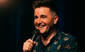 A man with dark hair and a beard smiles into a microphone, wearing a patterned shirt.