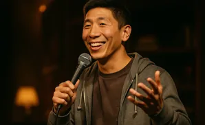 A man tells a joke into a microphone, smiling and gesturing with his hands.
