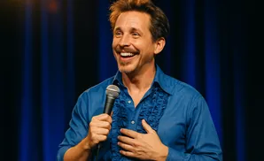 A smiling man in a blue shirt with ruffled front holds a microphone and his other hand on his chest.