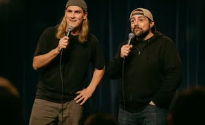Two men in baseball caps and casual clothes stand on a stage holding microphones, seemingly performing.