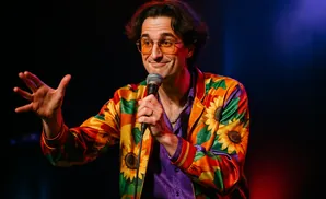 A man with dark hair and orange glasses holds a microphone and gestures with his left hand, wearing a colorful jacket with sunflowers.