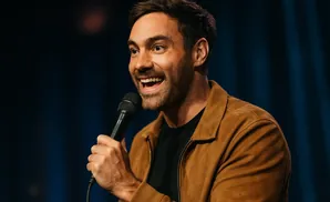 A man in a brown jacket holds a microphone and laughs.