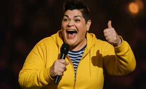 A woman in a yellow hoodie holds a microphone and gives a thumbs-up, smiling widely.