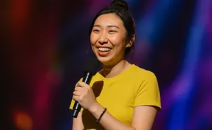 A woman with her hair in a bun, wearing a yellow shirt, laughs while holding a microphone on a colorful stage.