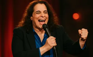 A comedian laughs into a microphone, her fist clenched in triumph, on a red-lit stage.
