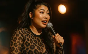 Woman in leopard print top holding a microphone and speaking into it on a dark stage with a spotlight.