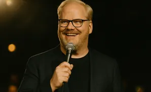 A man with blonde hair and glasses, wearing a black suit, smiles while holding a microphone.