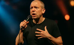 A man with a microphone and an expressive face gestures with his hand during a performance.