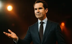 A man in a dark suit and brown tie gestures with his open hand, looking to the side.