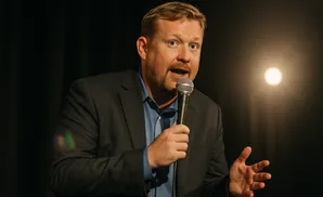 A man in a suit holds a microphone and speaks, gesturing with his hand, under a spotlight.