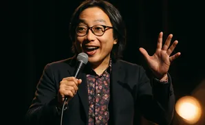 A man with glasses holds a microphone and raises his hand, smiling on a dark stage.