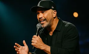 A man in a black baseball cap and shirt holds a microphone and gestures with his left hand while speaking.