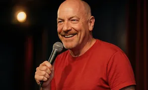 A smiling bald man in a red t-shirt holds a microphone onstage.