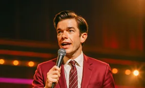 Comedian John Mulaney in a red suit speaking into a microphone on stage.