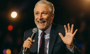 Jon Stewart smiles and holds a microphone, gesturing with his other hand.