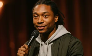 A Black man with dreadlocks holds a microphone while wearing a gray hoodie and a dark jacket.