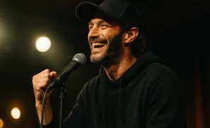 A man in a black hoodie and baseball cap laughs into a microphone, fist clenched.