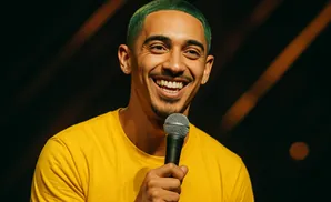A smiling man with green hair and a yellow shirt holds a microphone.