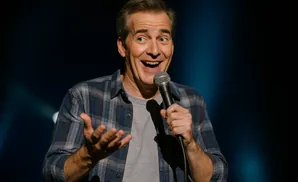 A man in a blue plaid shirt smiles and gestures with his hand while holding a microphone.