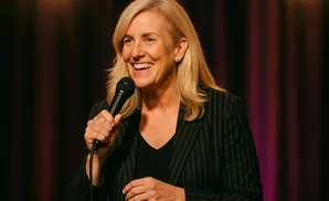 A blonde woman in a pinstripe jacket smiles while holding a microphone.