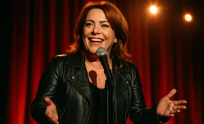 A woman with red hair in a black leather jacket laughs into a microphone on a stage with red curtains.