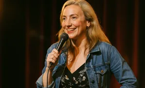A woman with blonde hair in a denim jacket and floral dress smiles as she holds a microphone.