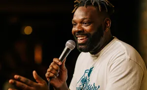 A man with dreadlocks and a beard smiles as he holds a microphone and speaks.