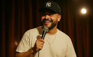 A man with a beard and baseball cap smiles while holding a microphone.