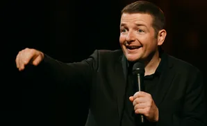 A man in a black suit smiles and gestures while holding a microphone.