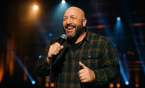 Bald comedian with a beard laughs while holding a microphone on stage with stage lights.