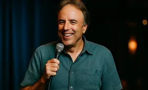 Comedian Kevin James holds a microphone and smiles broadly on stage in a blue shirt.