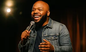 A Black man wearing a denim jacket laughs while holding a microphone on stage.